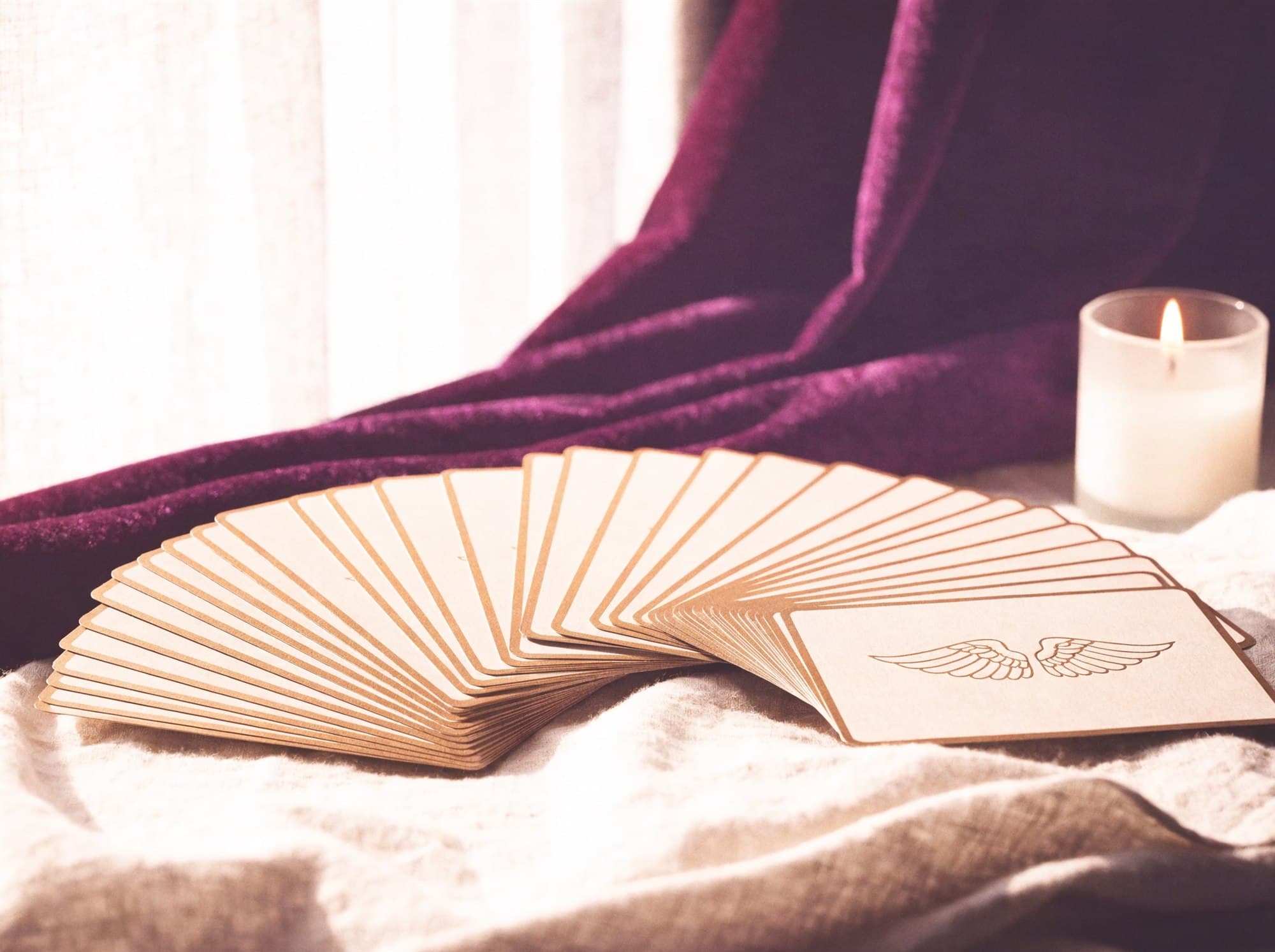 A gilt-edged angel oracle deck fanned on ivory linen beside a candle and plum velvet